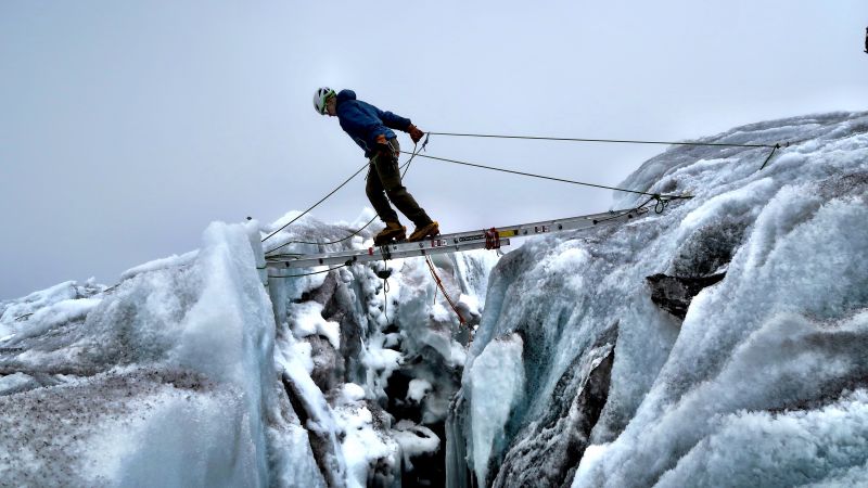 A climber crosses between a glacier crevasse on a ladder, using two ropes for support.