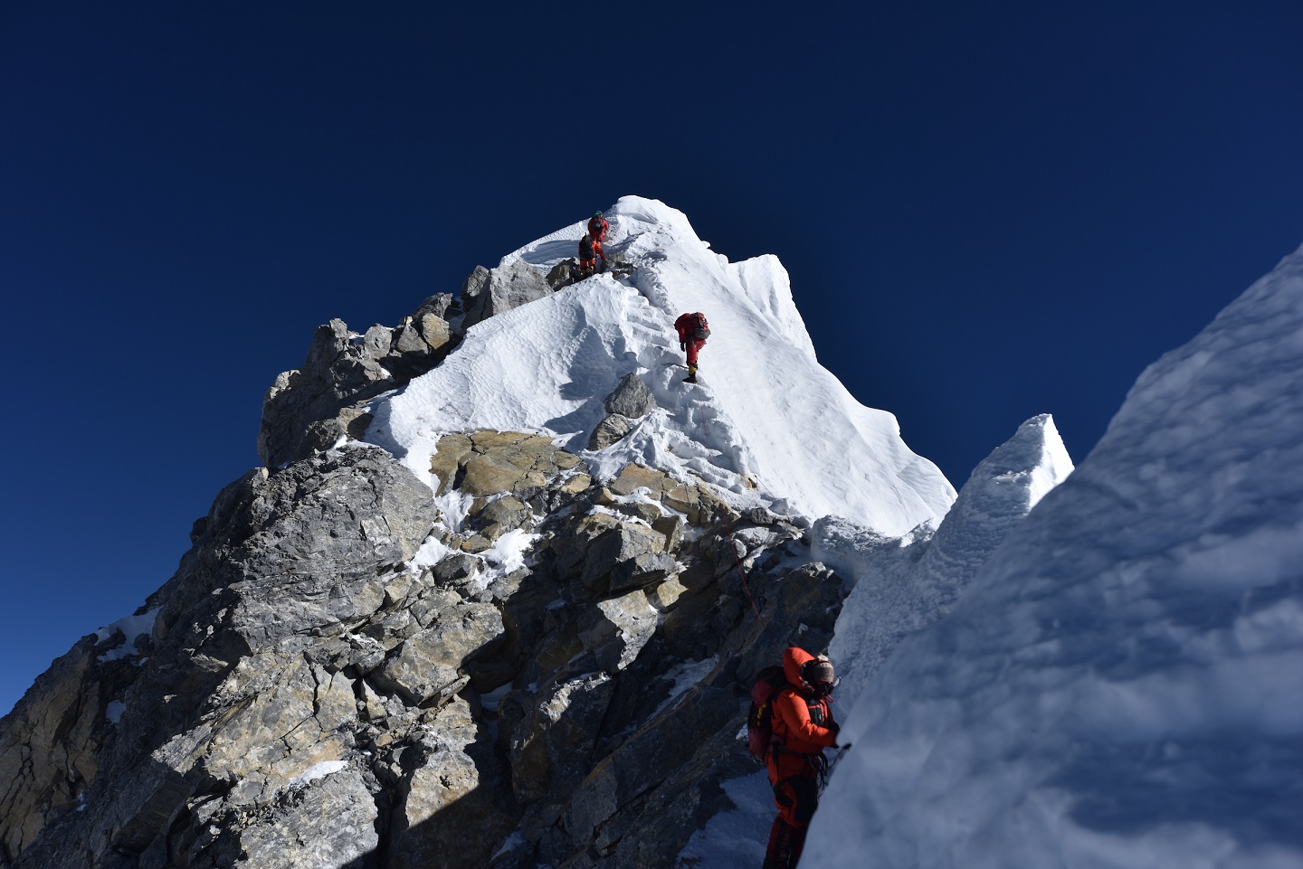 A photo of four climbers ascending a snowy mountain top in the Himalayas. Each climber is dressed in red down jackets and pants. The mountain is Mount Everest.