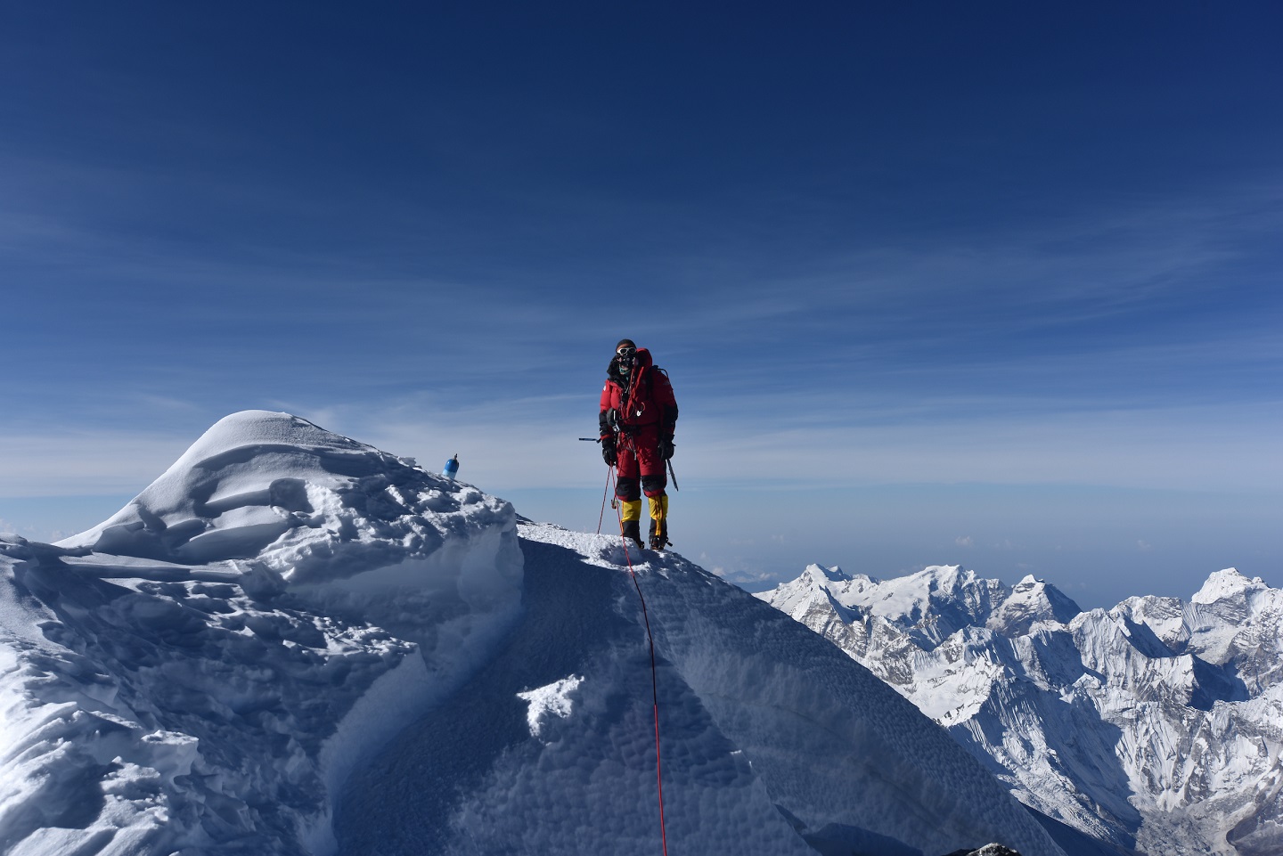 A lone climber stands on top of a snowy mountain peak. The peak is Mount Everest. The climber is dressed in red and using supplemental oxygen. In the background are the Himalayas.