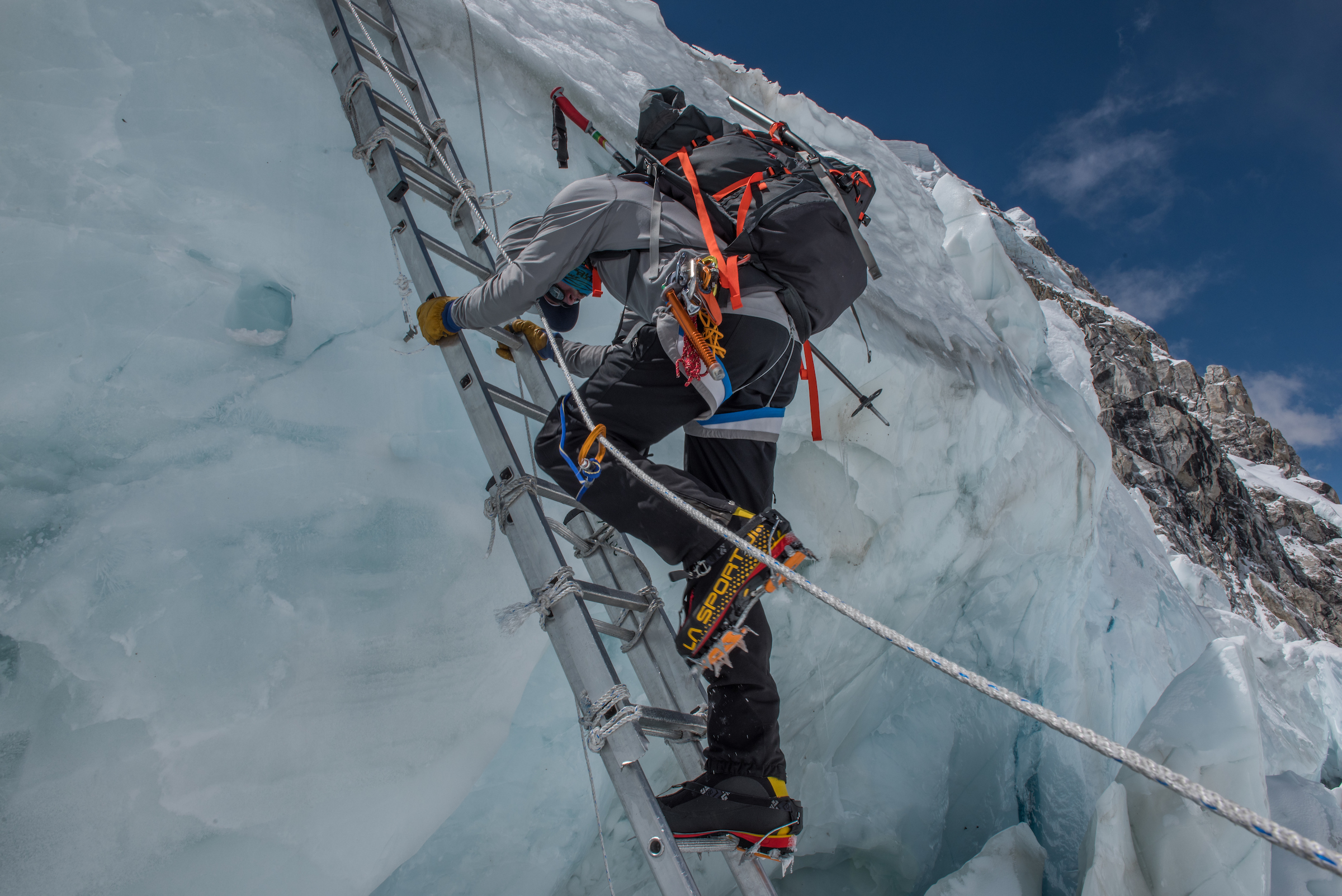 A man climbing up a ladder on a bergschrund. He is attached to a rope with full winter mountaineering gear including a backpack, ice axe, ice screws, etc.