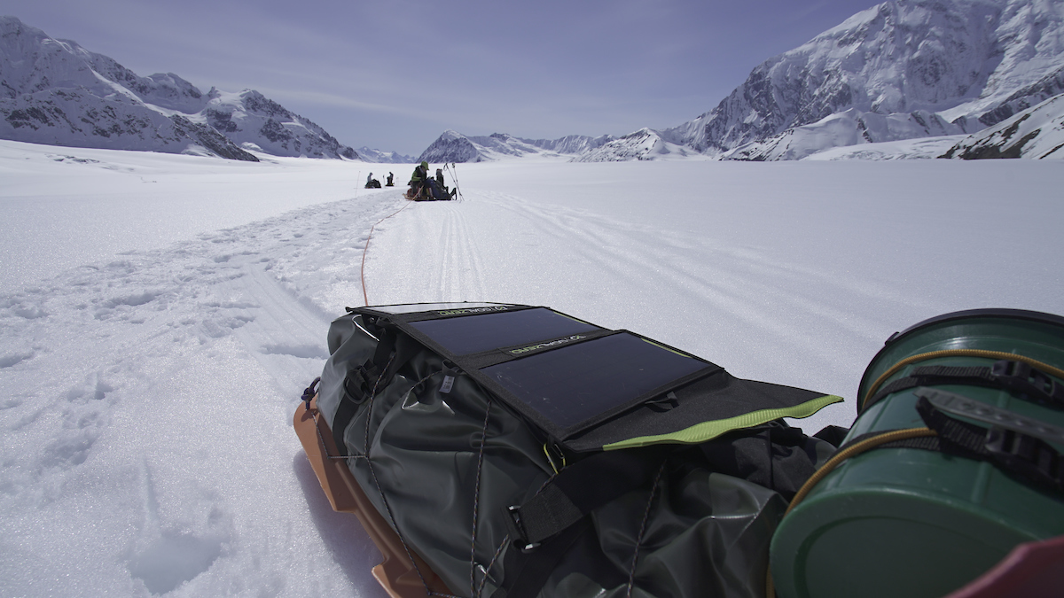A gear sled up close on a flat glacier with snow on top. In the background are more team members and mountains on both sides of the glacier valley. 