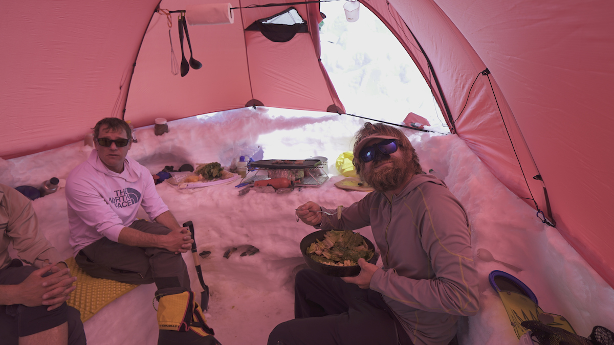Two men sit in a mountaineering kitchen tent. Both are facing the camera and one is smiling holding a bowl of salad. 