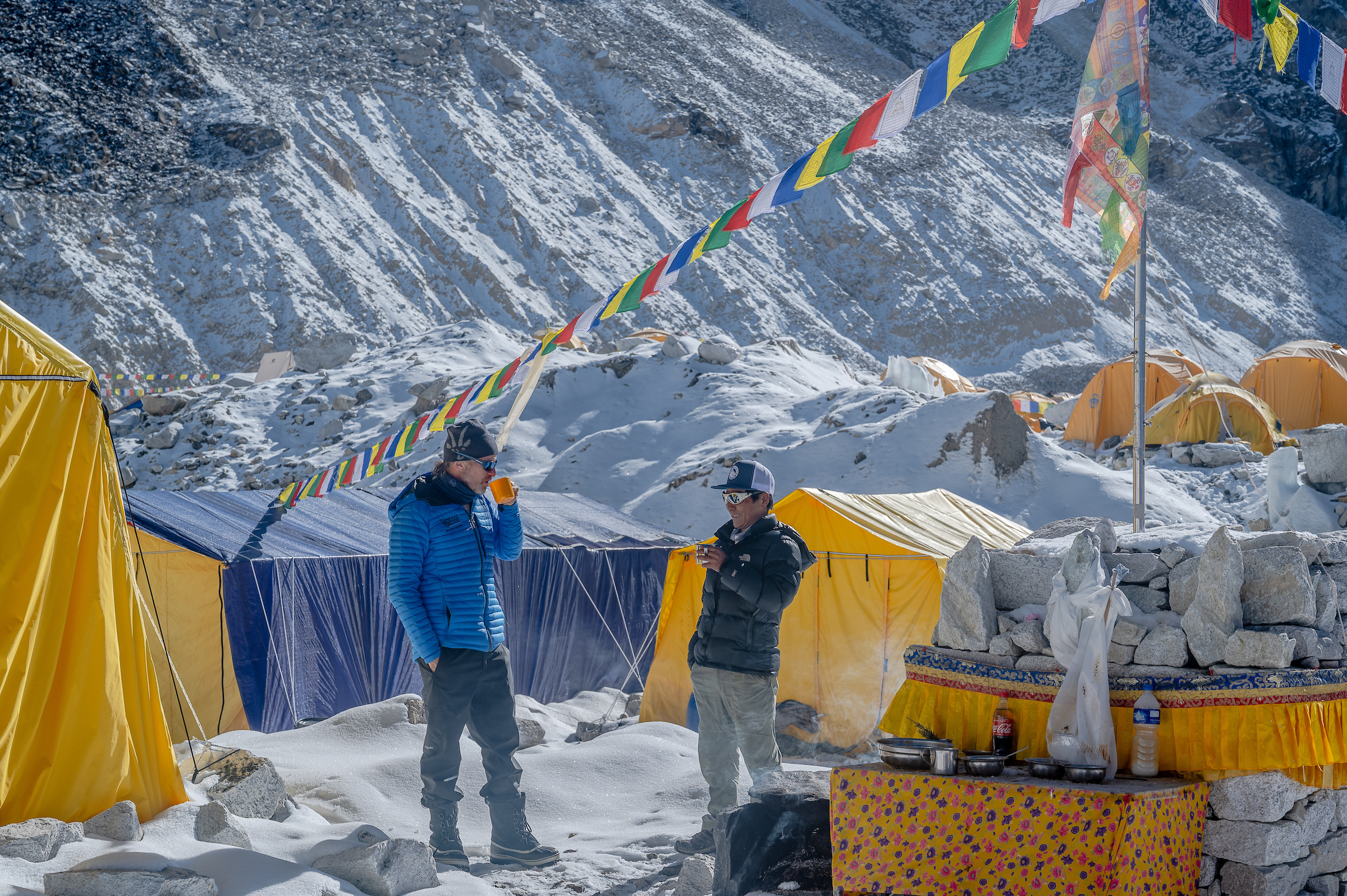 Two men are standing in the middle of Everest base camp with one sipping on a hot drink.