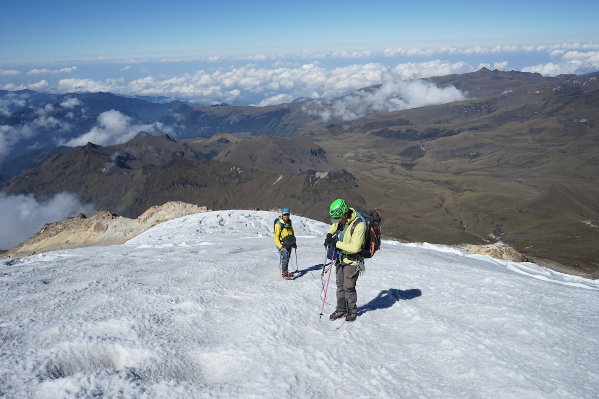 Two alpinists are standing in ahigh altitude snowfield with brown and far reaching mountains in the background.